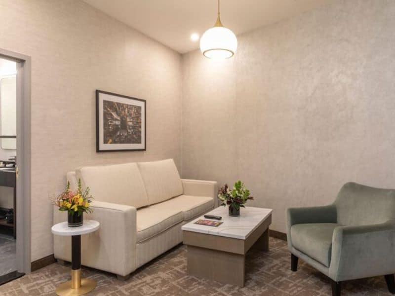 Stylish hotel room at Broadway Plaza Hotel featuring a cozy seating area with a white sofa, green chair, decorative flowers, and contemporary art on the wall, highlighting modern amenities in the Nomad district.