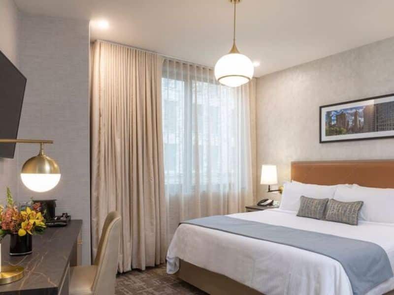 Elegant hotel room at Broadway Plaza Hotel featuring a king bed, contemporary decor, flat-screen TV, and work desk, with natural light from large window and stylish lighting fixtures.