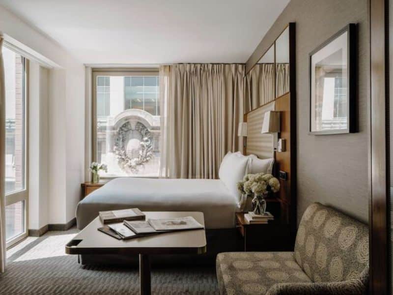 Elegant hotel room at Club Quarters Hotel Grand Central, New York, featuring a cozy bed, modern furnishings, natural light from large windows, and a small seating area with a table, emphasizing comfort and style for business and leisure travelers.