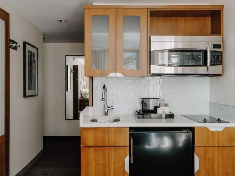 Kitchenette area in Club Quarters Hotel Grand Central featuring wooden cabinets, stainless steel microwave, sink, and black refrigerator, highlighting the hotel's amenities for business and leisure travelers.