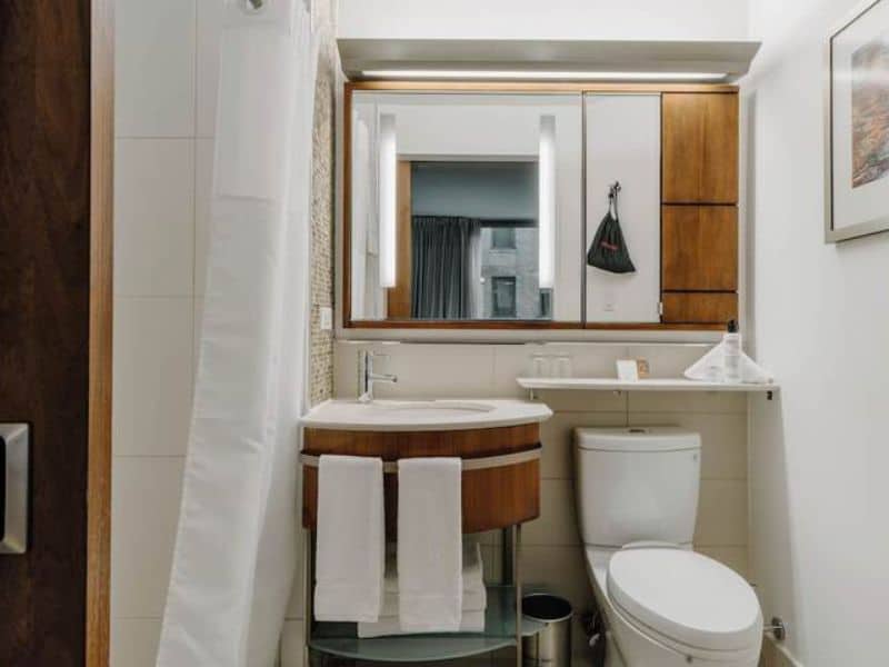 Modern hotel bathroom with circular vanity, mirror, and toiletries; features white towels, a toilet, and stylish wooden cabinetry, reflecting the amenities of Club Quarters Hotel Grand Central, New York.