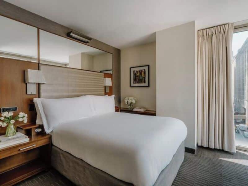 Elegant hotel room at Club Quarters Hotel Grand Central, featuring a queen bed, bedside lamps, modern decor, and large windows with city views.