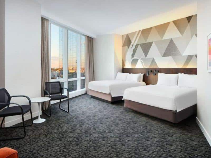 Modern hotel room at Courtyard Long Island City featuring two queen beds, floor-to-ceiling windows, contemporary decor, and seating area, highlighting views of the New York City skyline.