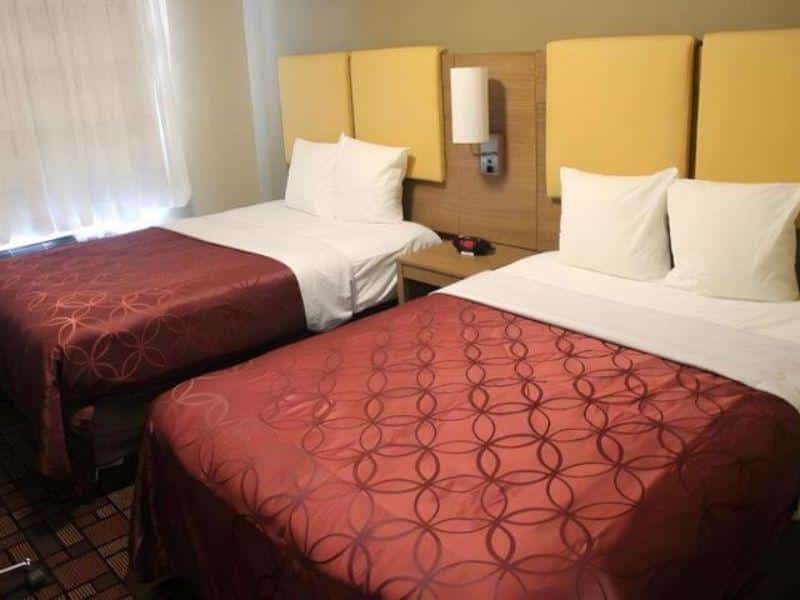 Cozy hotel room at Days Inn by Wyndham Brooklyn Borough Park featuring two beds with red patterned bedspreads, yellow headboards, and natural light from a nearby window.