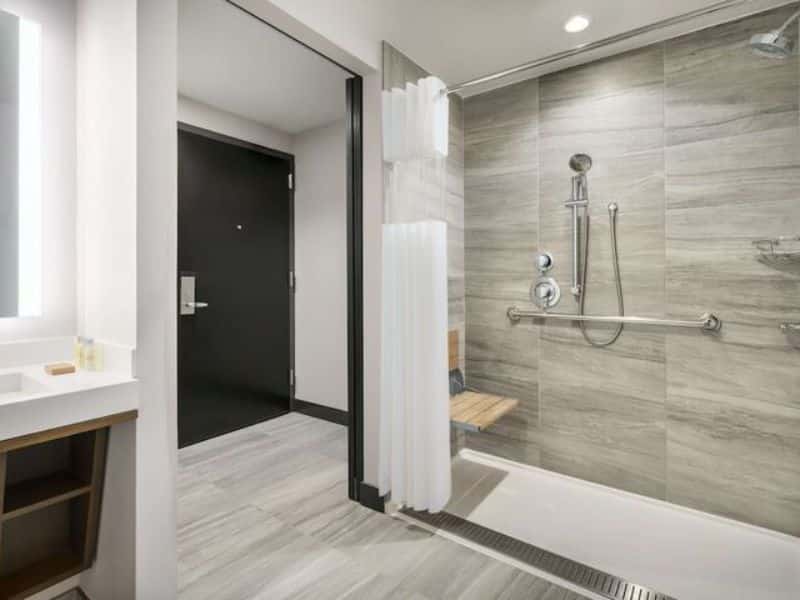 Accessible hotel bathroom featuring a walk-in shower with grab bars, a wooden bench, and a modern sink area, emphasizing the DoubleTree by Hilton New York Times Square South's commitment to guest comfort and accessibility.