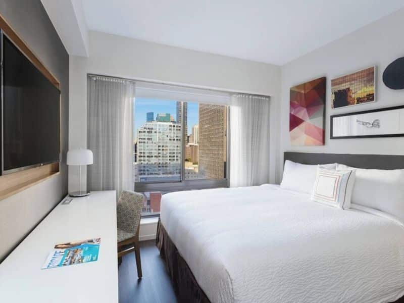 Cozy hotel room at Fairfield Inn & Suites by Marriott New York Manhattan/Central Park, featuring a king-size bed, flat-screen TV, desk, and large window with city views.