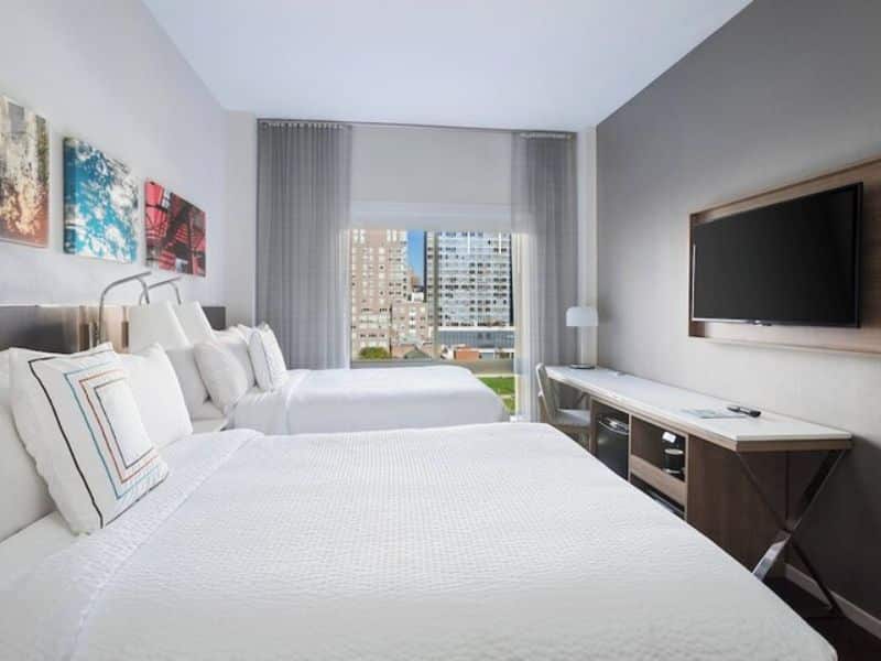 Cozy guest room at Fairfield Inn & Suites by Marriott New York Manhattan/Central Park, featuring two beds, flat-screen TV, modern decor, and city views through large window.