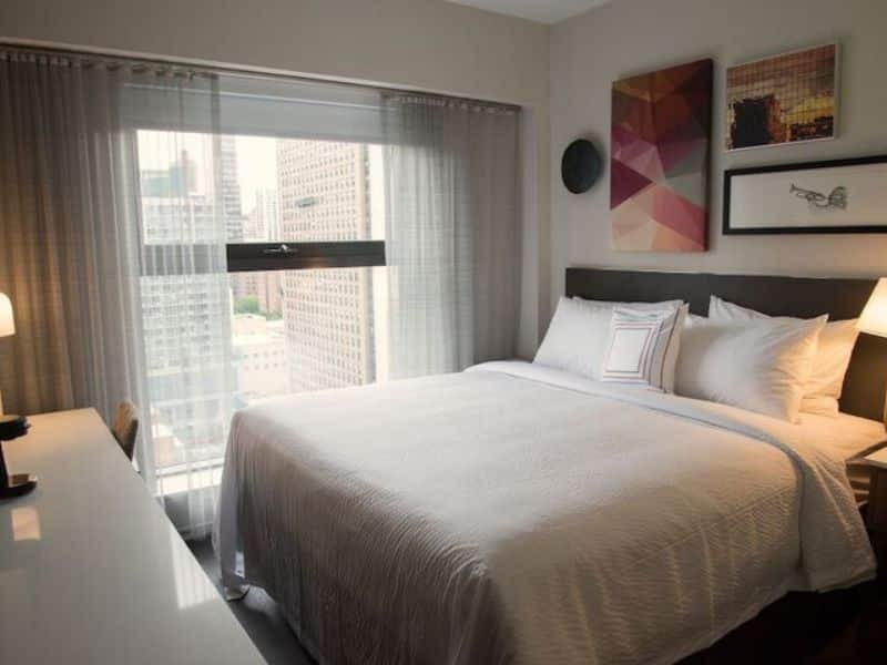 Cozy hotel room at Fairfield Inn & Suites by Marriott New York Manhattan/Central Park, featuring a queen bed, city views through large windows, modern decor, and a desk area with a lamp.