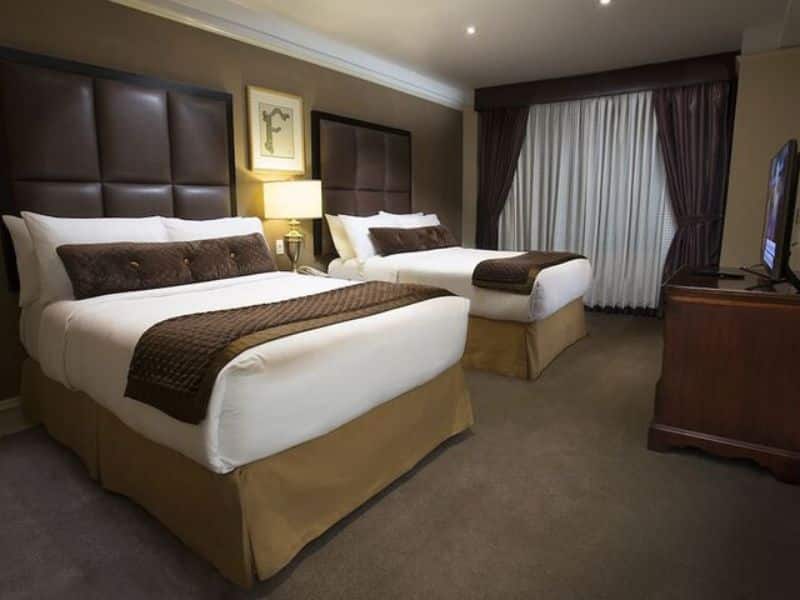 Modern hotel room at Fitzpatrick Manhattan featuring two queen beds with brown and white bedding, leather headboards, a wooden dresser with a TV, and natural light from a window with curtains.