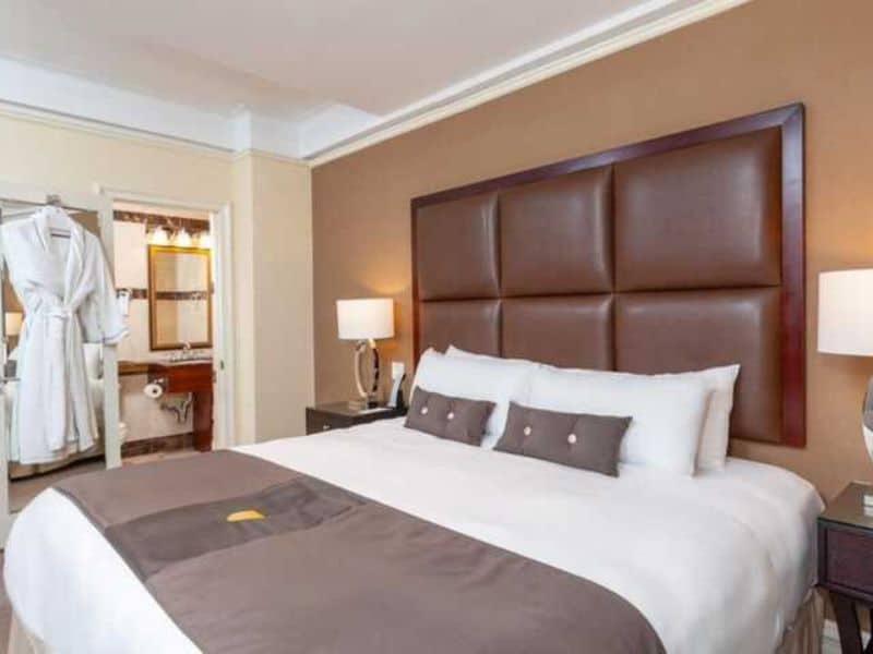 Modern hotel room at Fitzpatrick Manhattan featuring a king-sized bed with decorative pillows, leather headboard, bedside lamps, and a view of an en-suite bathroom with a robe hanging nearby.