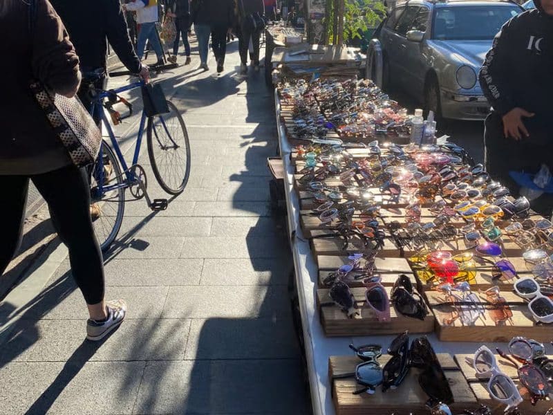 Person walking past a vibrant outdoor market stall displaying an array of colorful sunglasses, with a cyclist nearby, capturing the lively atmosphere of Williamsburg's eclectic markets.