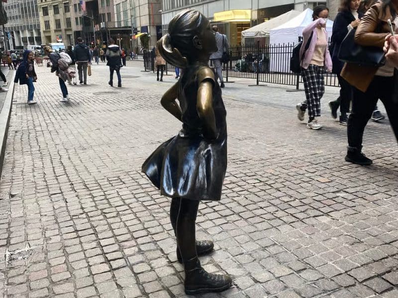 Bronze statue of a girl in a dress standing confidently on a cobblestone street in Southern Manhattan, surrounded by pedestrians, symbolizing empowerment and resilience in a historic neighborhood.