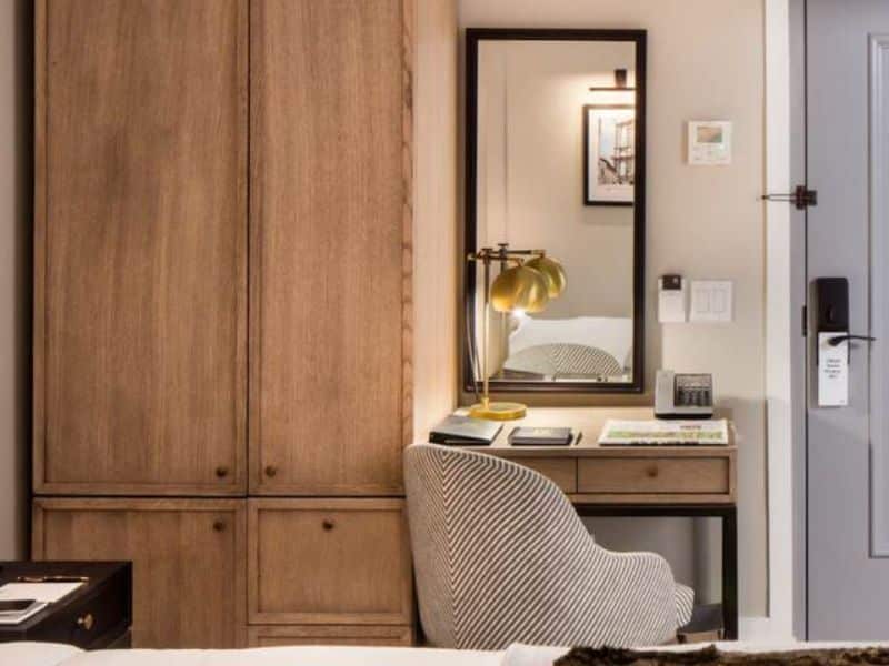 Cozy hotel room corner with wooden wardrobe, desk featuring a lamp and mirror, and a striped chair, highlighting the HGU New York's boutique style and amenities.