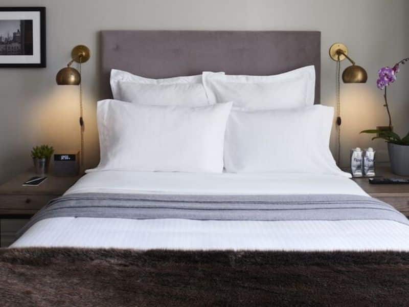Cozy hotel room at HGU New York featuring a plush bed with white linens, decorative pillows, bedside lamps, and modern amenities, highlighting luxury and comfort in Midtown Manhattan.