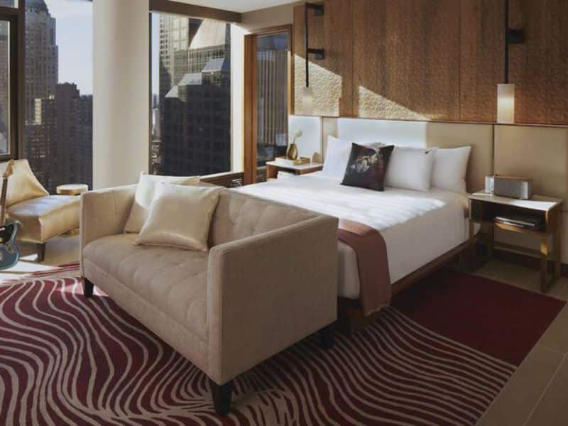 Stylish hotel room at Hard Rock Hotel New York featuring a queen bed, modern furnishings, a cozy sofa, large windows with city views, and a vibrant area rug.