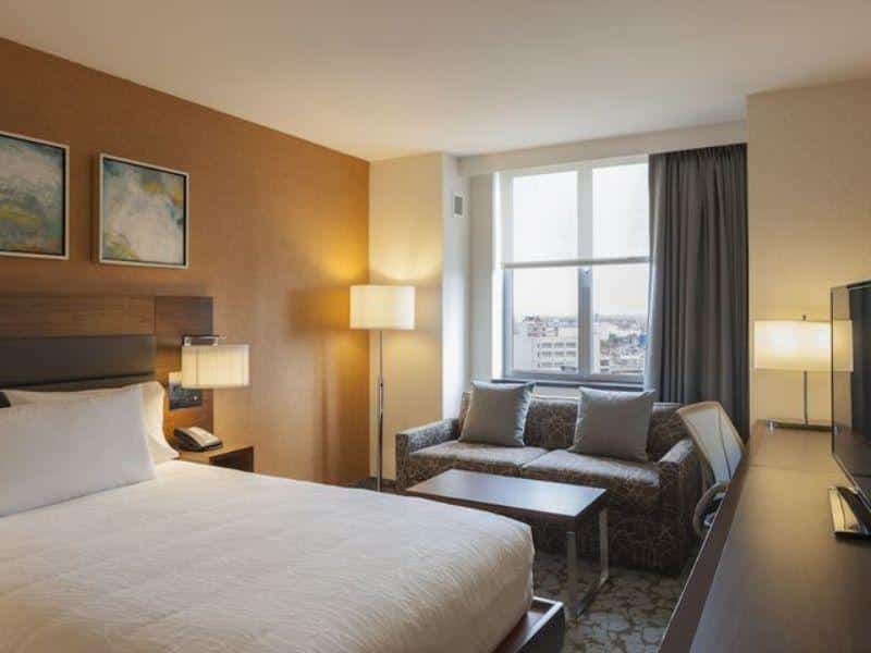 Cozy hotel room at Hilton Garden Inn Long Island City with a king-sized bed, modern furnishings, a sofa, and large windows showcasing city views, emphasizing comfort and convenience for travelers.