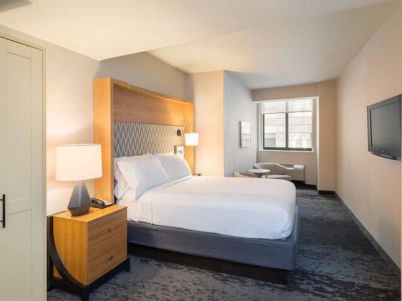 Modern hotel room at Holiday Inn New York City – Wall Street featuring a queen bed, bedside lamp, flat-screen TV, and seating area near a window.