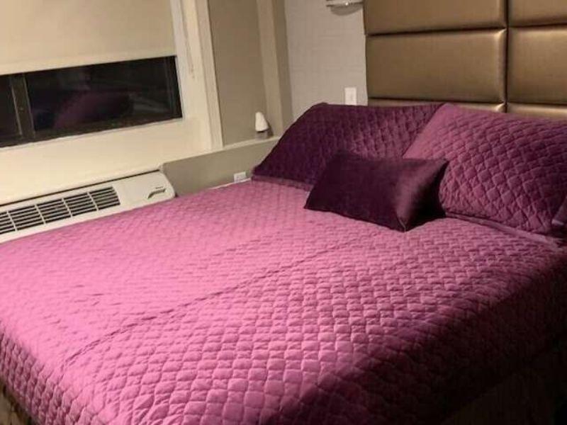 Cozy hotel room featuring a purple quilted bedspread, accent pillows, and a window, highlighting comfortable accommodations near LaGuardia Airport.
