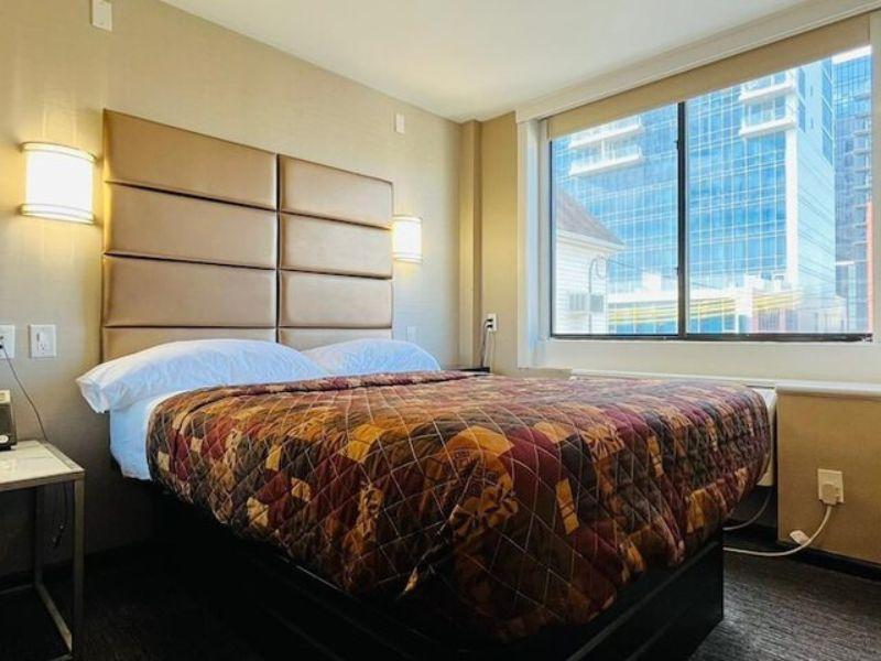 Cozy hotel room featuring a patterned bedspread, bedside table, and large window with city view, highlighting comfortable accommodations near LaGuardia Airport.