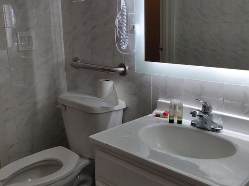 Bathroom with a toilet, sink, and illuminated mirror, featuring toiletries and grab bars, showcasing amenities at Hotel Key in Flushing, NY.