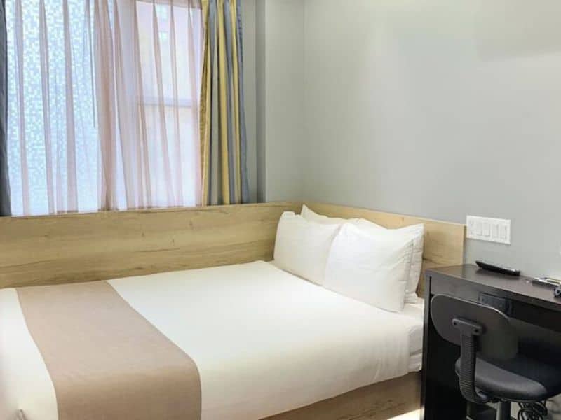 Cozy hotel room at Madison LES Hotel featuring a queen bed, wooden accents, desk area, and natural light from window drapes.