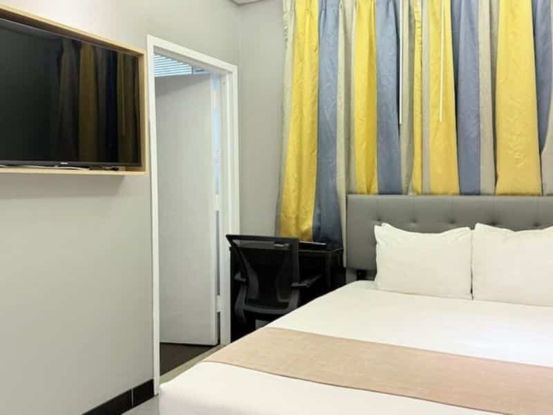 Cozy hotel room at Madison LES Hotel featuring a queen bed, flat-screen TV, and colorful drapes, with a desk and door leading to an adjoining space.