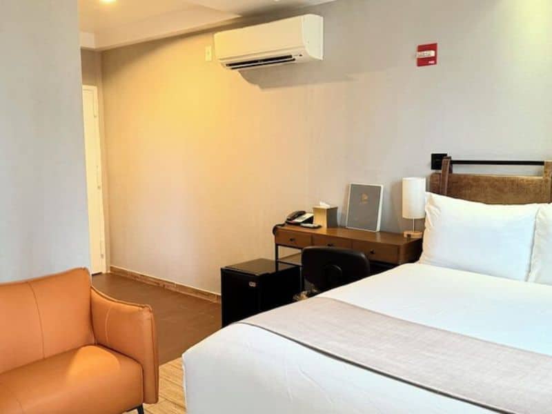 Hotel room at Madison LES Hotel featuring a queen bed, orange accent chair, desk with lamp, and air conditioning unit, showcasing modern decor and comfortable amenities.