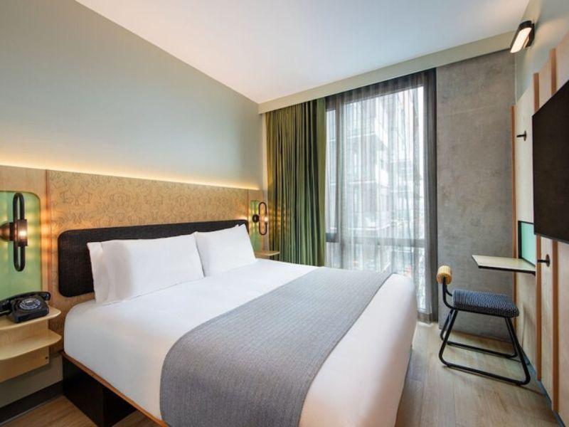 Stylish hotel room at Moxy Brooklyn Williamsburg featuring a queen bed, floor-to-ceiling windows, modern decor, and a workspace, highlighting the vibrant atmosphere and amenities offered.