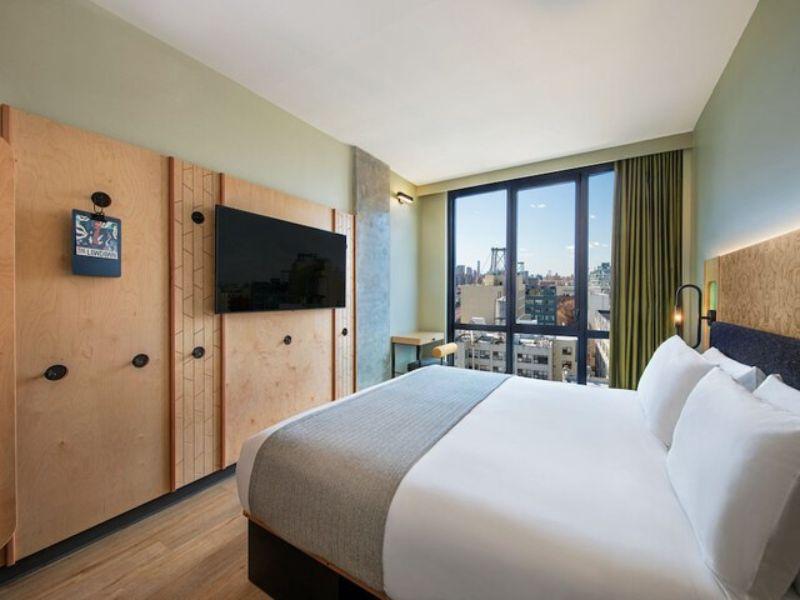 Stylish hotel room at Moxy Brooklyn Williamsburg featuring a queen bed, floor-to-ceiling windows with city views, modern decor, and a flat-screen TV.