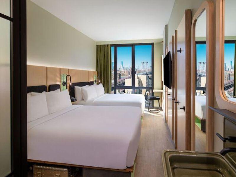 Stylish hotel room at Moxy Brooklyn Williamsburg with two beds, floor-to-ceiling windows, and city views, featuring modern decor and amenities.