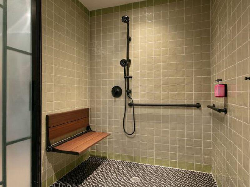 Accessible hotel shower with grab bars, a wooden bench, and modern fixtures in Moxy Brooklyn Williamsburg.