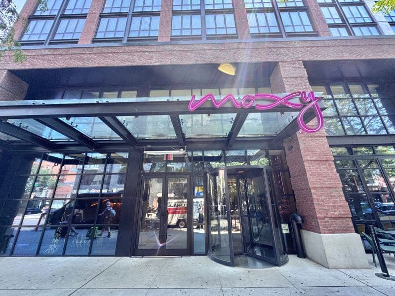 Moxy Brooklyn Williamsburg hotel entrance with modern glass façade and vibrant pink signage, showcasing a welcoming atmosphere in a lively neighborhood.
