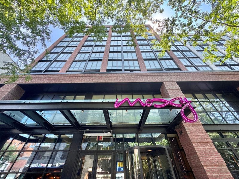 Moxy Brooklyn Williamsburg hotel exterior with vibrant pink signage, modern architecture, and lush greenery framing the entrance.