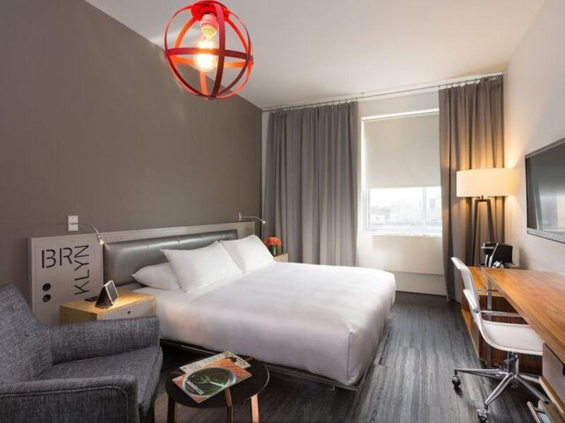 Stylish hotel room at NU Hotel Brooklyn featuring a modern design, king-size bed, gray walls, cozy seating area, and desk, with natural light from large window.