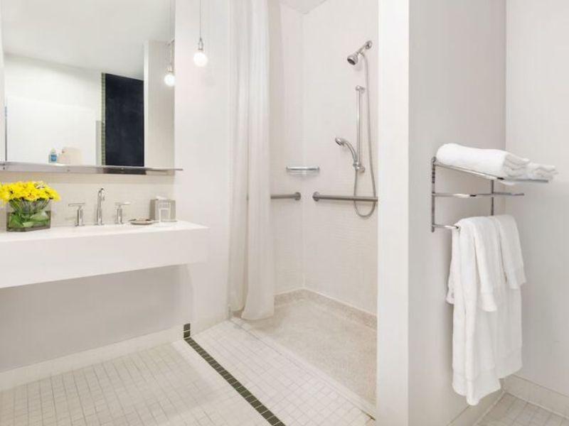 Modern hotel bathroom featuring a spacious shower area, stylish sink with dual faucets, decorative flowers, and neatly arranged towels, reflecting the amenities of NU Hotel Brooklyn.