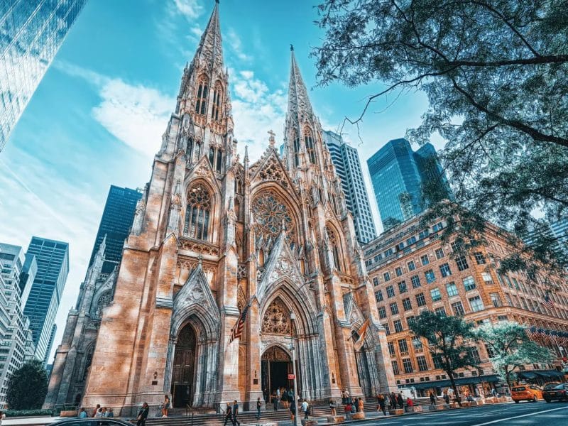 St. Patrick&rsquo;s Cathedral in New York City, iconic filming location from The Godfather, showcasing Gothic architecture amidst skyscrapers, with pedestrians and vehicles in the foreground.