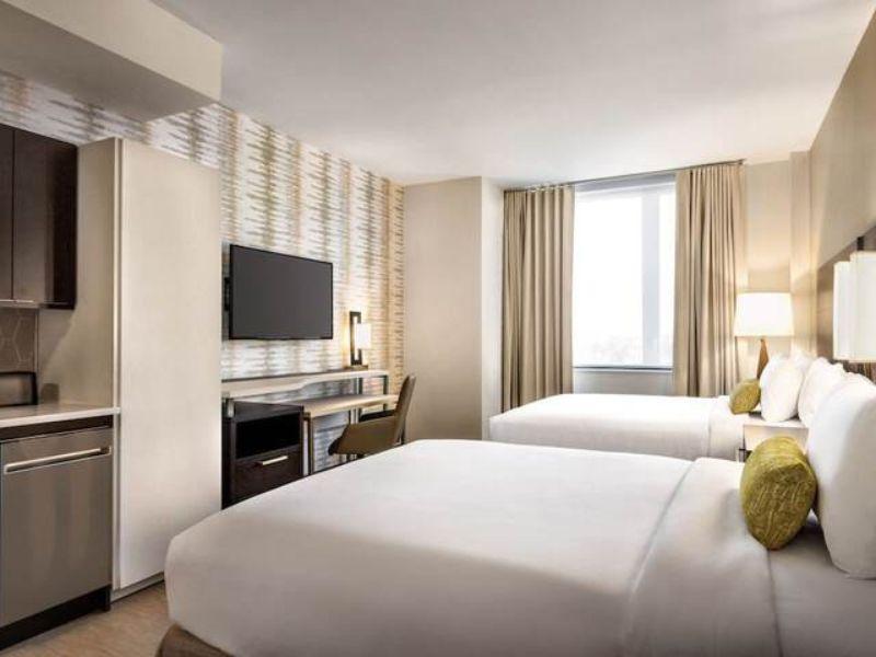 Modern hotel room with two queen beds, kitchenette, desk, and large window, highlighting amenities at Residence Inn by Marriott New York JFK Airport.