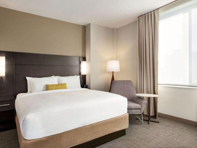 Modern hotel room at Residence Inn by Marriott New York JFK Airport with a queen bed, cozy seating area, and large window, highlighting comfortable amenities for travelers.