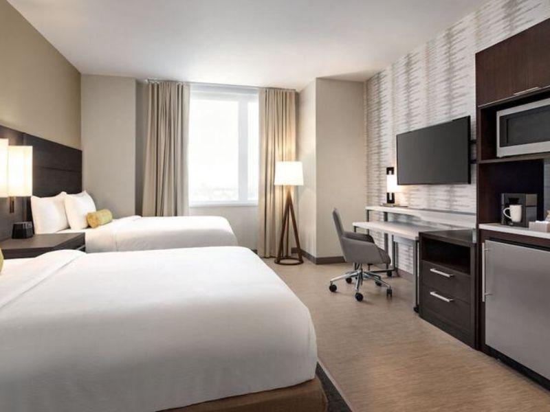 Modern hotel room with two beds, desk, TV, kitchenette, and natural light, highlighting amenities at Residence Inn by Marriott New York JFK Airport.