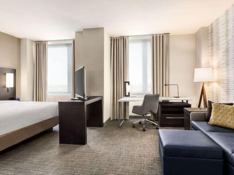 Modern hotel room at Residence Inn by Marriott New York JFK Airport, featuring a king-sized bed, workspace with desk and chair, flat-screen TV, and seating area with sofa, large windows with natural light, and contemporary decor.