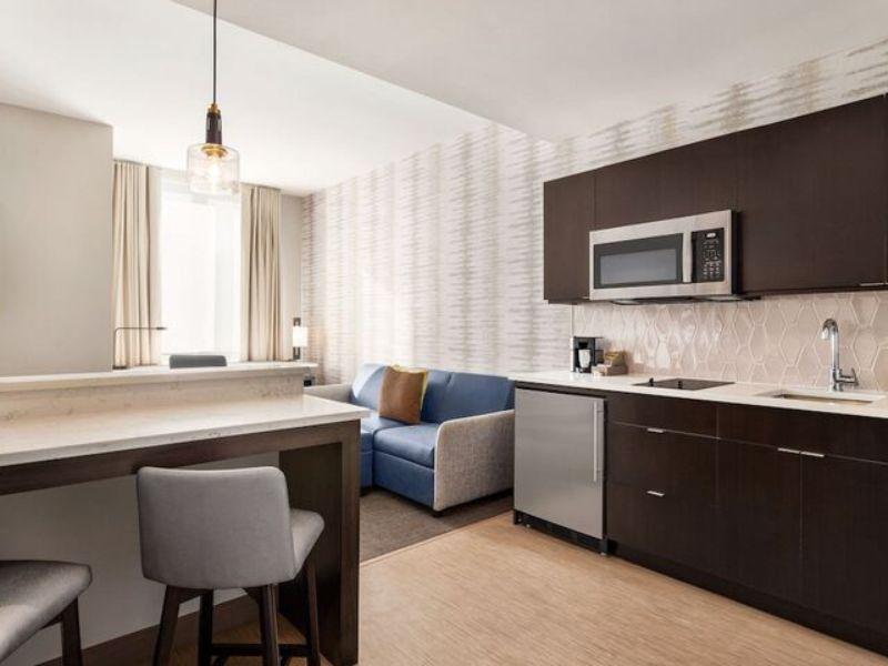 Modern hotel suite at Residence Inn by Marriott New York JFK Airport, featuring kitchenette, dining area, and comfortable seating with natural light.