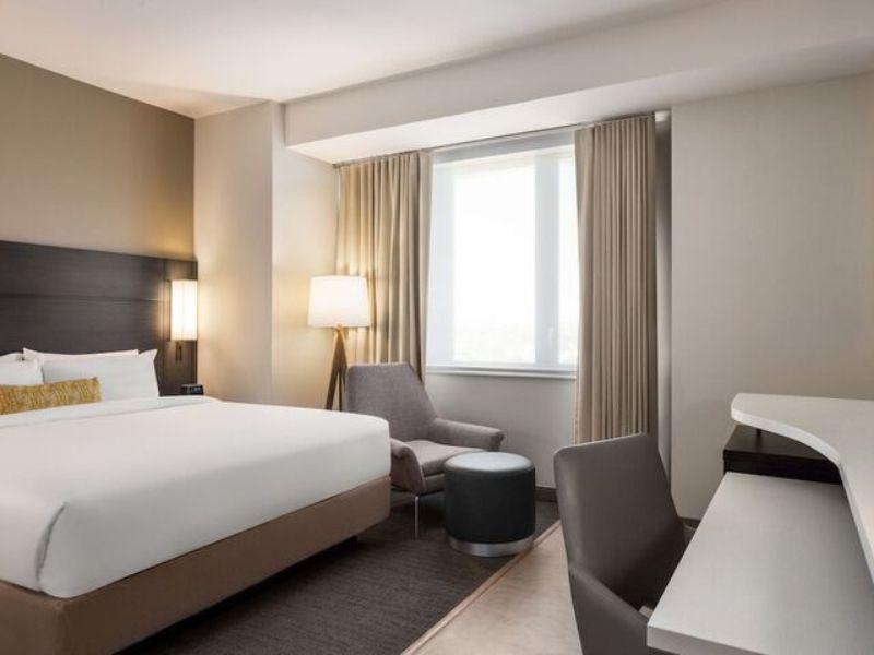 Modern hotel room at Residence Inn by Marriott New York JFK Airport, featuring a queen bed, seating area with armchair, desk, and large window with natural light.