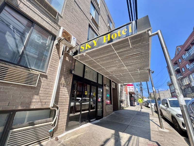 Sky Hotel entrance featuring a large sign, located in Flushing, near LaGuardia Airport, with a clear blue sky above and nearby street activity.