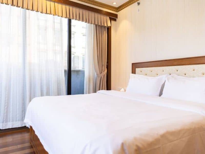 Elegant hotel room with a king-sized bed, floor-to-ceiling windows draped with sheer curtains, and warm wooden flooring, showcasing modern comforts and city views at The Allen Hotel in New York City.