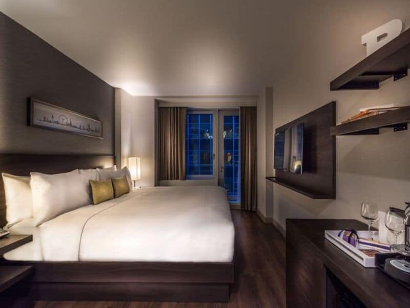 Stylish hotel room at The Bernic Hotel New York City, featuring a king-sized bed, floor-to-ceiling windows, modern decor, and a wall-mounted TV, highlighting amenities like Nespresso machine and complimentary Wi-Fi.