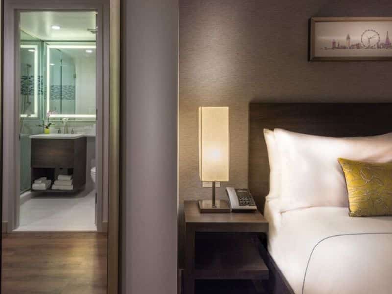 Stylish hotel room at The Bernic Hotel New York City, featuring a bedside lamp, phone, and a view of the modern bathroom with glass shower and amenities, highlighting the contemporary design and comfort offered by the Tapestry Collection by Hilton.