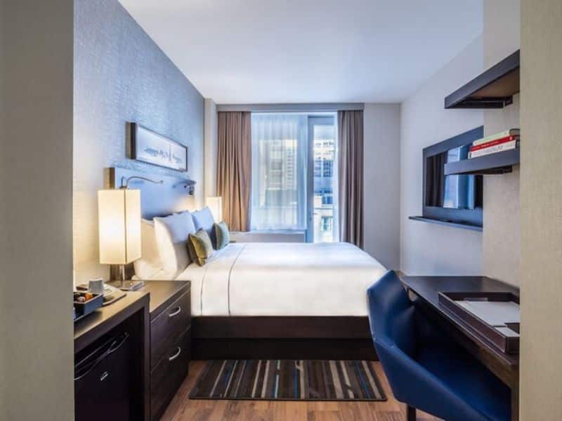 Stylish hotel room at The Bernic Hotel New York City, featuring a queen bed with decorative pillows, floor-to-ceiling windows, a work desk, and modern decor, emphasizing comfort and convenience for guests.