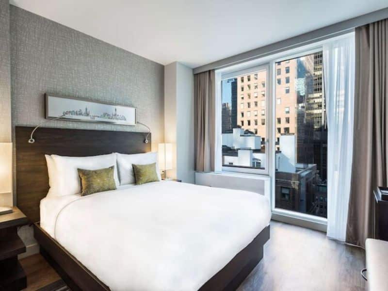 Stylish hotel room at The Bernic Hotel New York City, featuring a queen bed, floor-to-ceiling windows, modern decor, and city views, emphasizing comfort and luxury in Midtown East.
