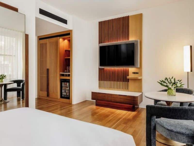 Stylish hotel room at The Bryant Park Hotel featuring hardwood floors, flat-screen TV, minibar, and modern decor with a small dining table and fresh flowers.