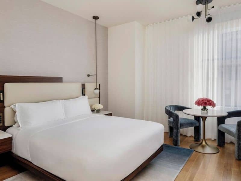 Modern hotel room at The Bryant Park Hotel featuring a queen-sized bed, stylish decor, hardwood floors, seating area with a round table, and natural light from large windows.